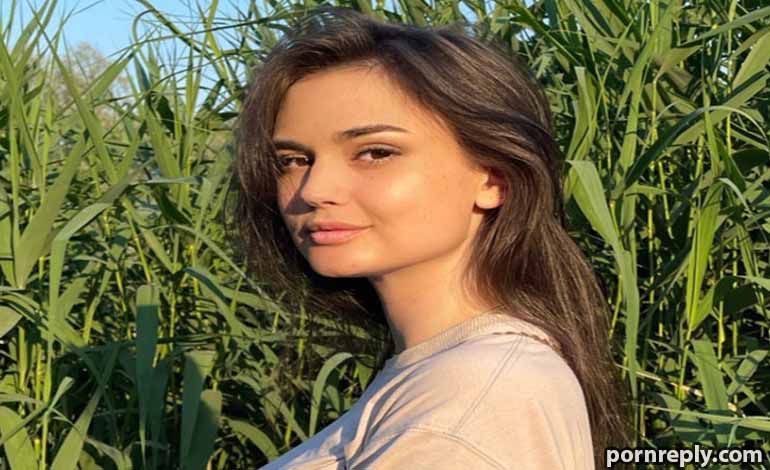 Hairy Cam Girls: How To Explore Fantasies Safely With Partner Side profile of a young woman with long brown hair standing in a field of tall green plants, sunlit and smiling slightly.