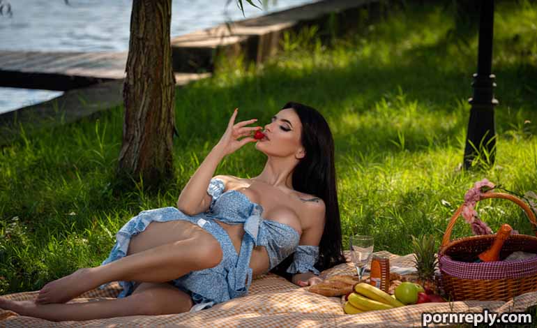 How a Furry Sex Doll Can Help Couples Talk and Connect Better Woman sunbathing on a checkered blanket by a lake, wearing a blue wrap top and biting a cherry; picnic basket and fruit nearby.